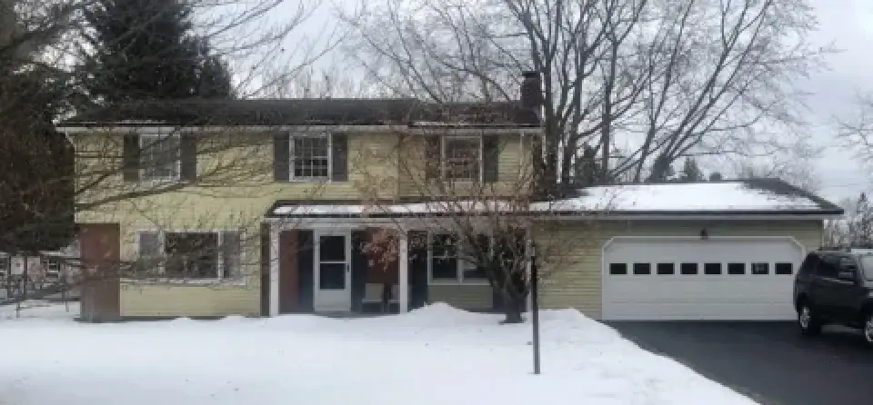 Two-story yellow house with attached garage, snowy lawn, bare trees, and a black SUV in driveway.