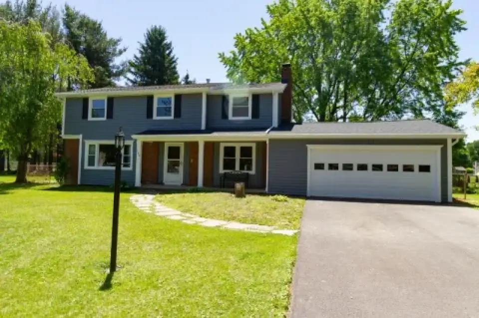 Two-story gray house with attached garage, green lawn, and trees on a sunny day.