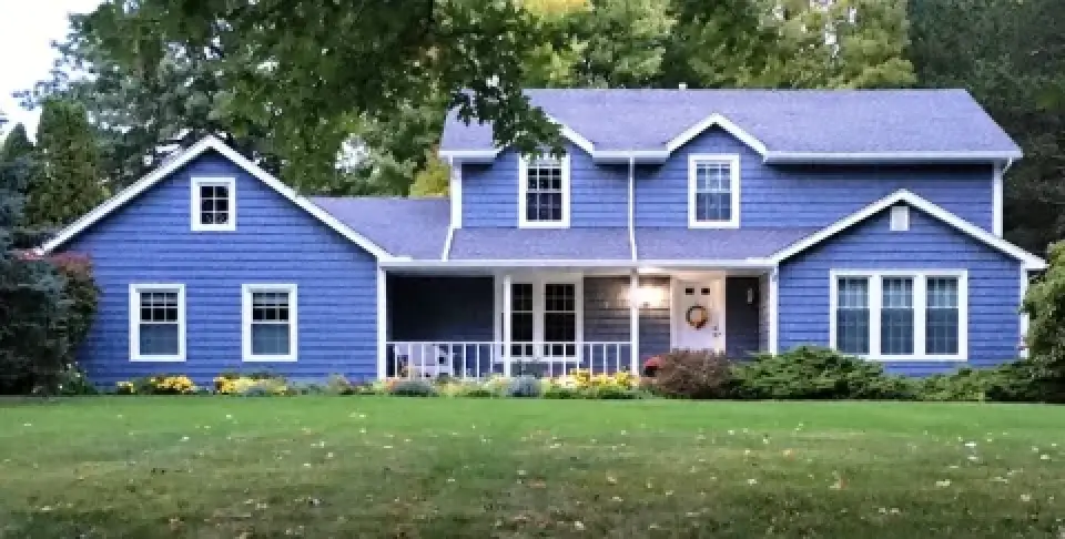 Blue house with white trim, front porch, and green lawn surrounded by trees and bushes.