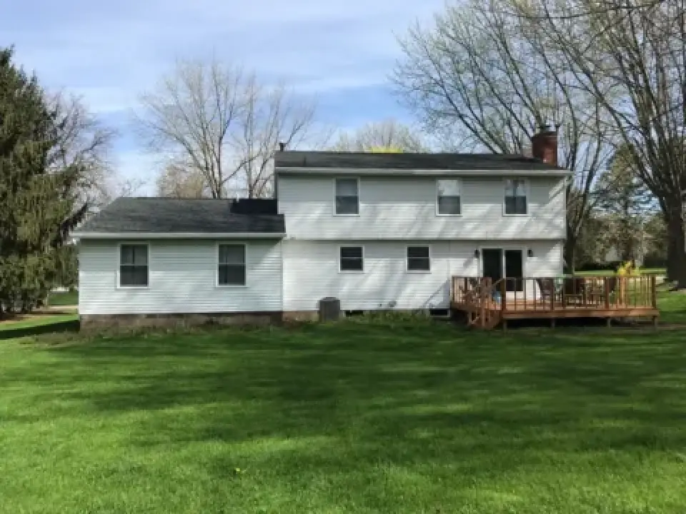 Two-story white house with a deck, large grassy yard, and trees in the background.