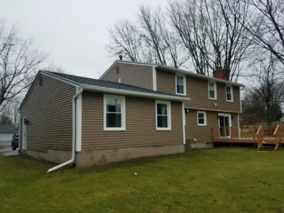 Two-story beige house with white trim, backyard lawn, deck, and leafless trees in the background.