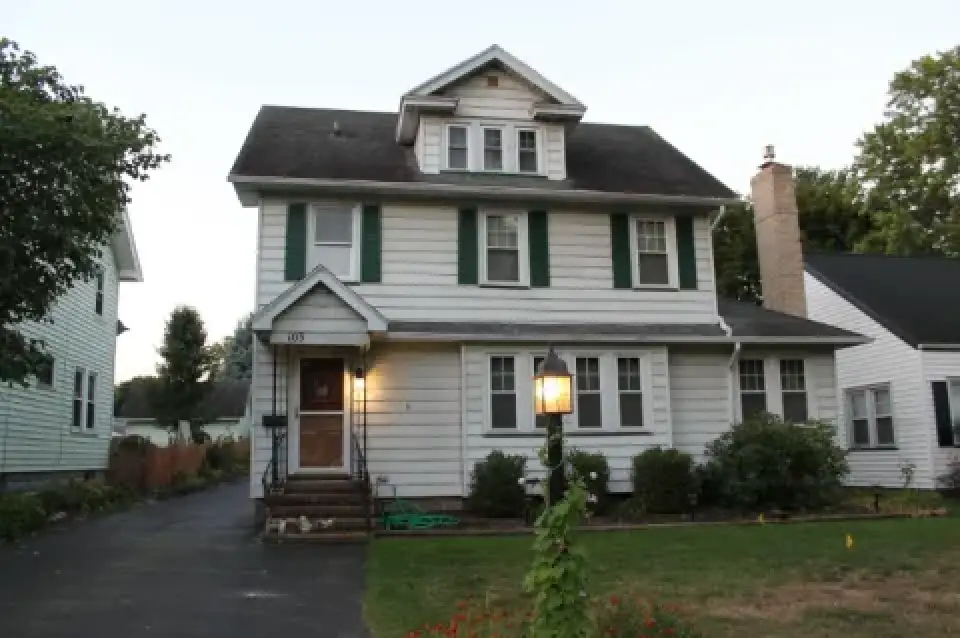 Two-story white house with green shutters, a front porch, and a lamp post in the yard.