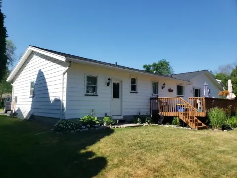 White single-story house with a wooden deck, surrounded by grass and plants on a sunny day.