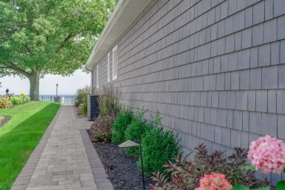 Gray-shingled house with a paved walkway, garden, and view of water in the background.