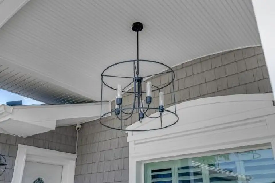 Black metal chandelier with candle-style lights hangs from a white beadboard ceiling outdoors.