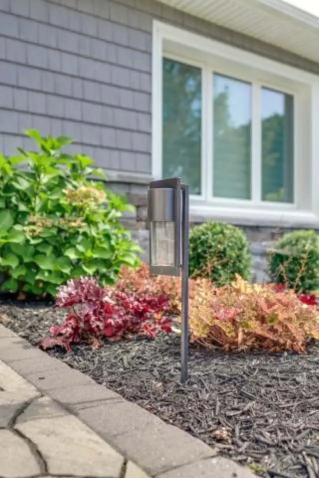 Modern garden light in landscaped flower bed near a house with shrubs and a large window.
