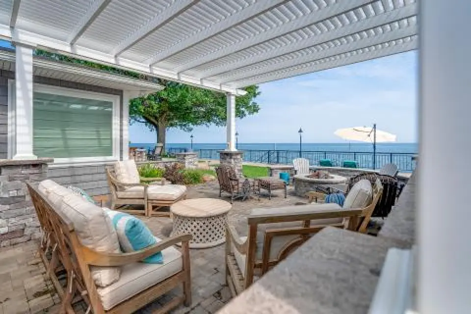 Patio with cushioned chairs and pergola, overlooking a lawn and water with seating and umbrella.
