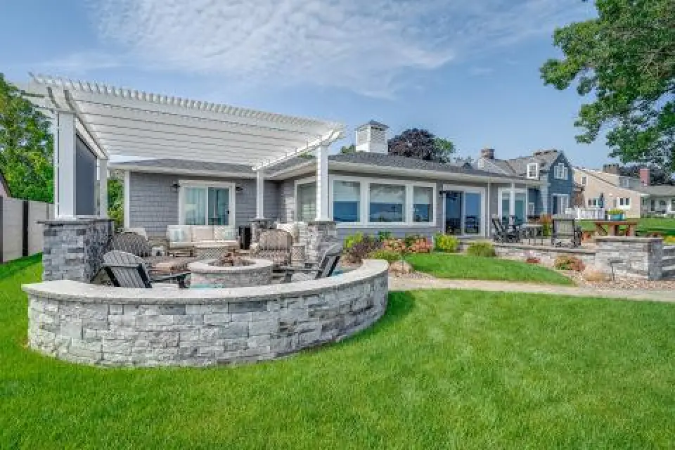 Single-story house with a pergola, stone patio, fire pit, and green lawn on a sunny day.