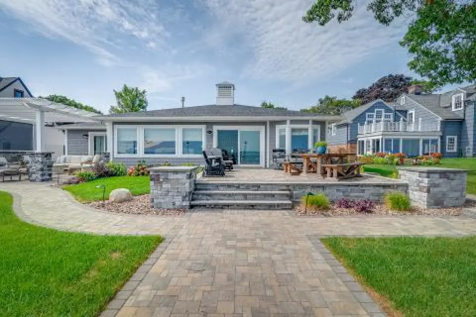 Single-story house with large windows, patio seating, and landscaped yard under a partly cloudy sky.