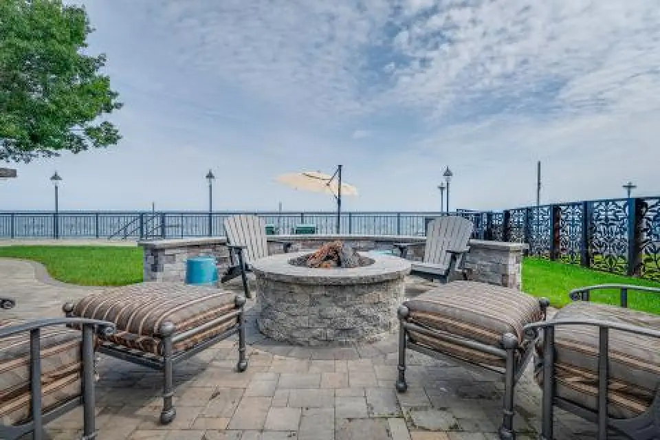 Outdoor fire pit with striped cushioned chairs, by a lake with a fence, umbrella, and cloudy sky.