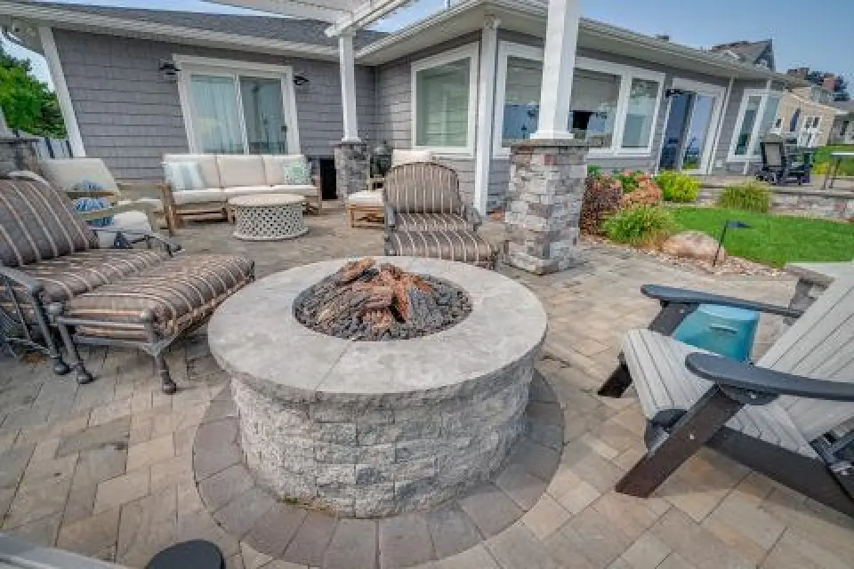 Outdoor patio with cushioned chairs around a stone fire pit, in front of a gray house.