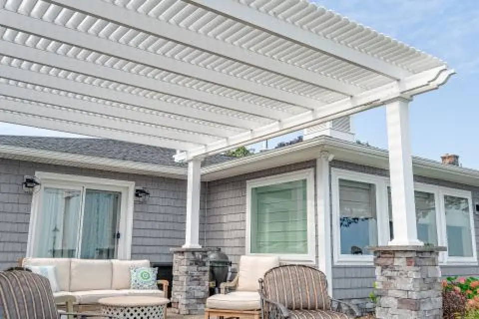 White pergola over a patio with outdoor seating in front of a gray house with large windows.