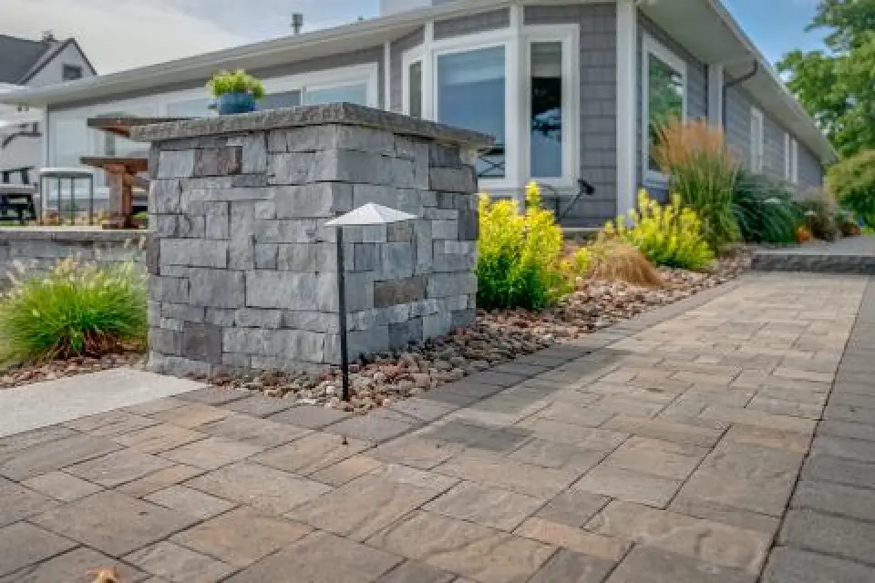 Stone pillar and garden light along a paved walkway beside a modern house with large windows.