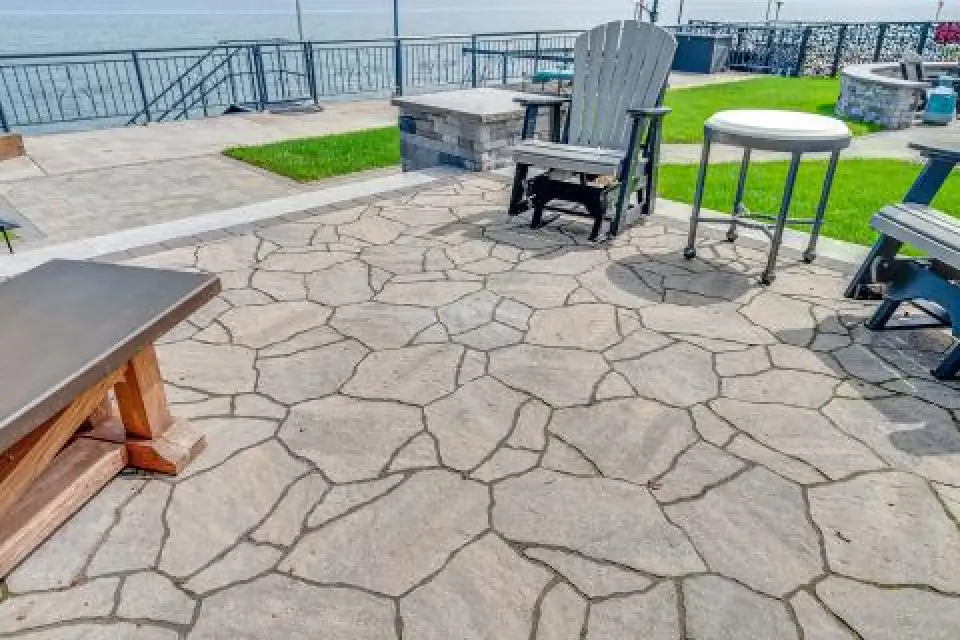 Stone patio with outdoor chairs and tables overlooking a body of water, with grass patches nearby.