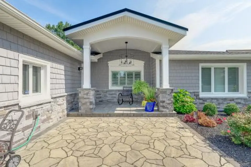 Front porch of a gray house with stone columns, a swing, plants, and a patterned walkway.
