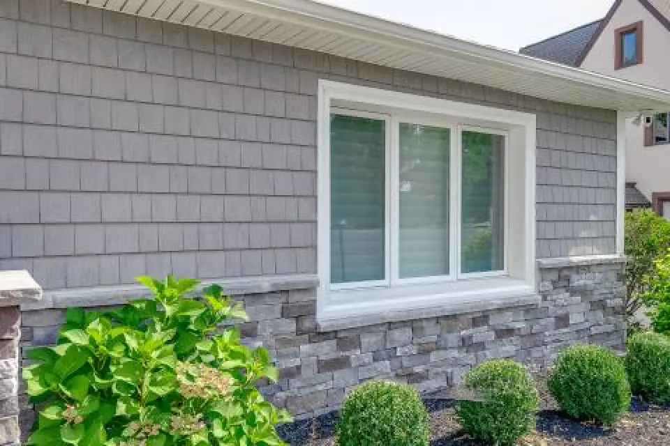 Gray house siding with white-trimmed window above stonework, shrubs planted in front.