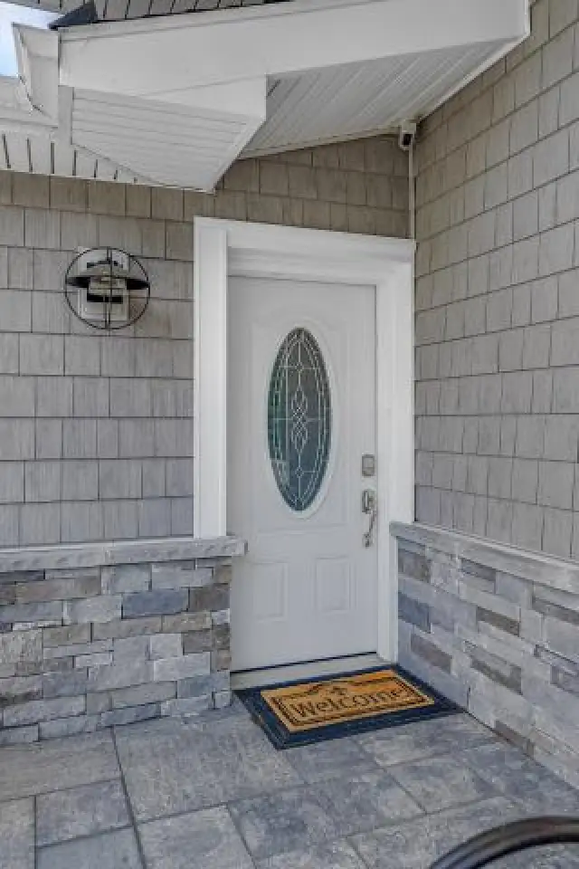White front door with oval window, welcome mat, stone wall, and outdoor light fixture above.