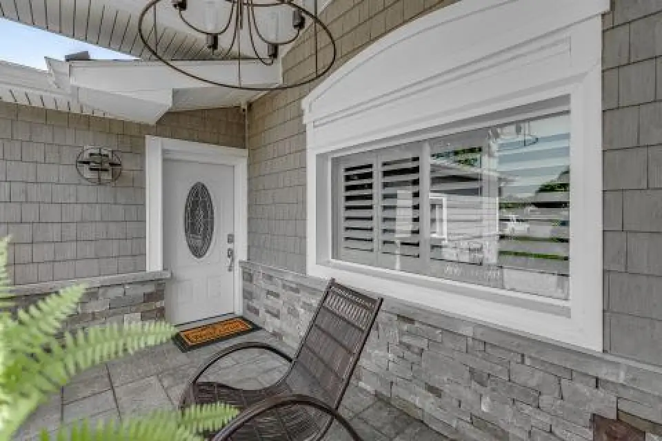 Covered porch with a white door, window with shutters, two chairs, and gray stone walls.