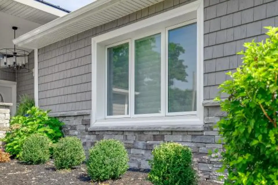A house window with gray siding, stone trim, and green shrubs in front of it.