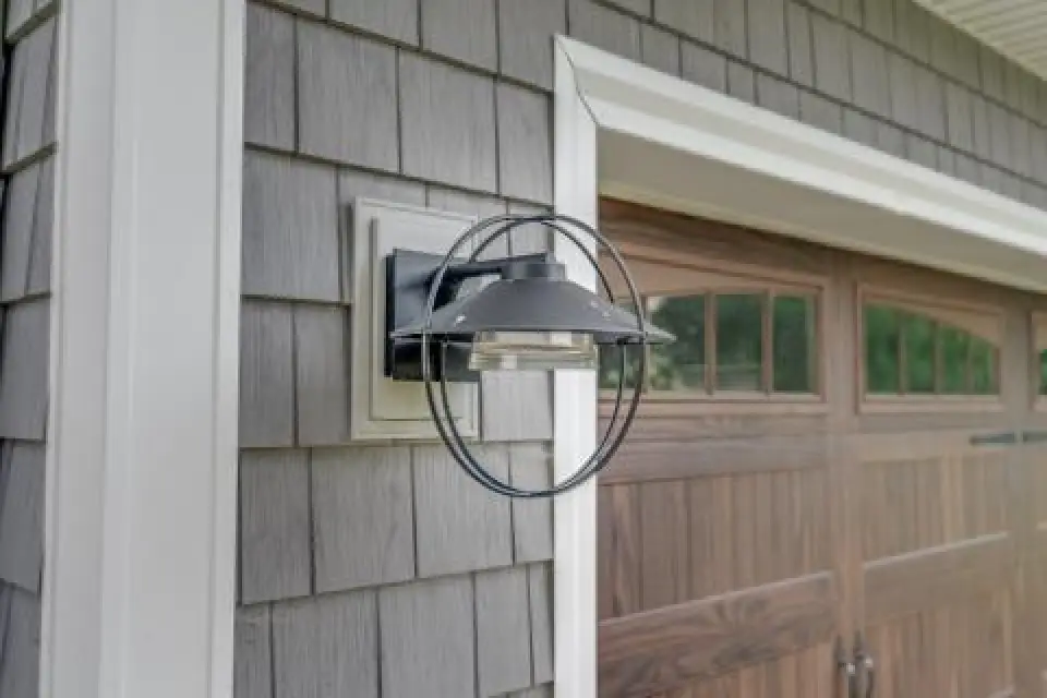Outdoor wall sconce light fixture mounted next to a wooden garage door on a gray house exterior.