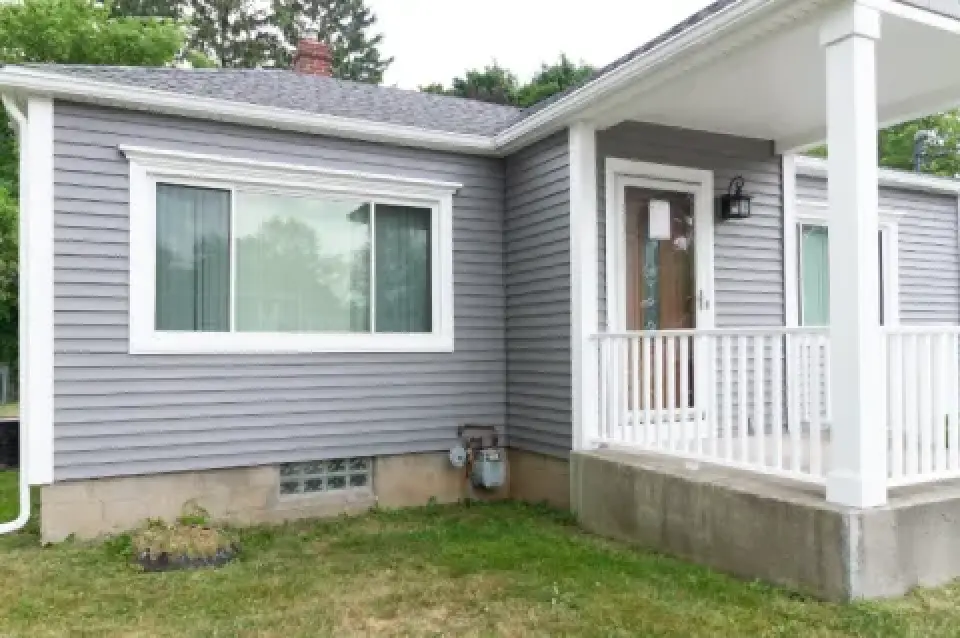 Gray house with white trim, a front porch, large window, and green grass lawn outside.
