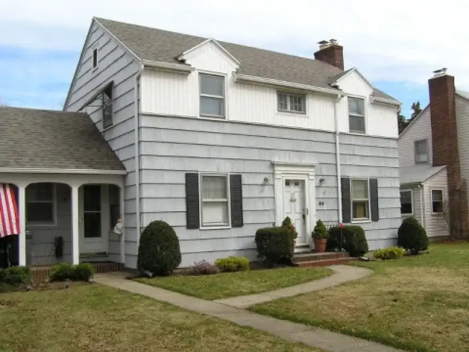 Light grey two-story suburban house with a front porch, shrubs, and a small lawn.