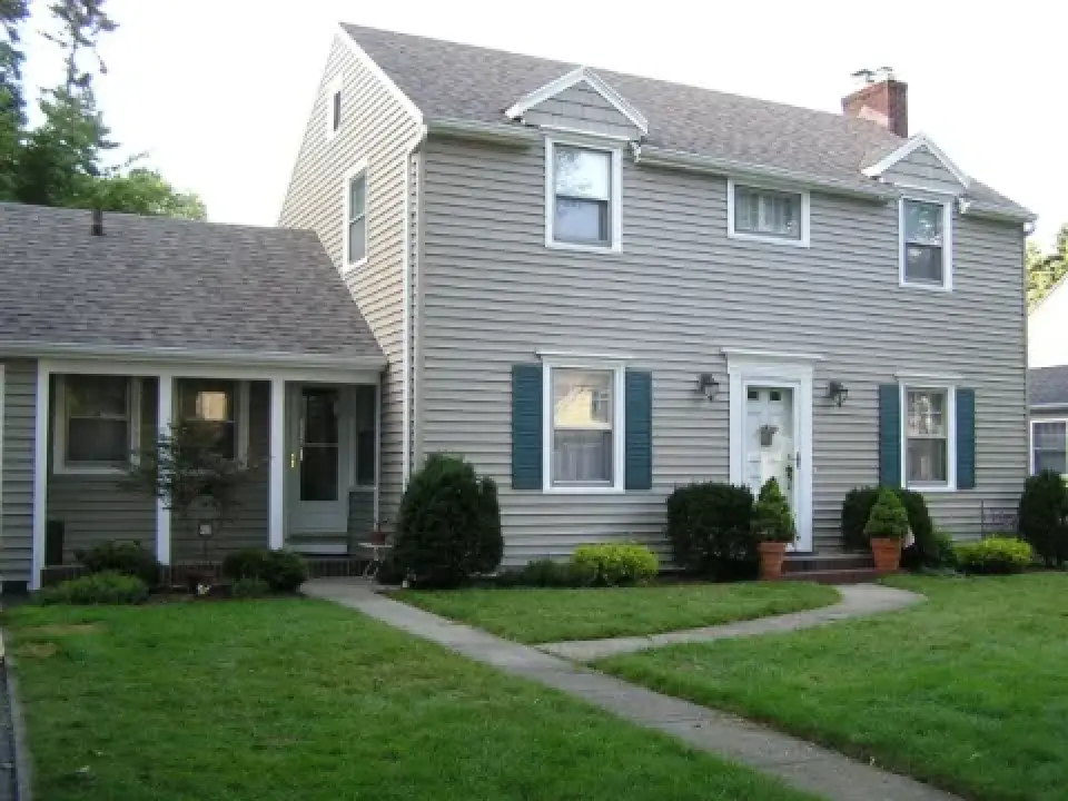 Two-story gray house with blue shutters, green lawn, bushes, and a paved walkway.