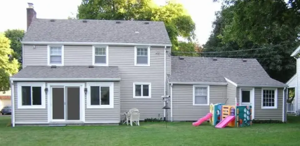 Two-story beige house with a playset, slide, and lawn chairs in a grassy backyard.