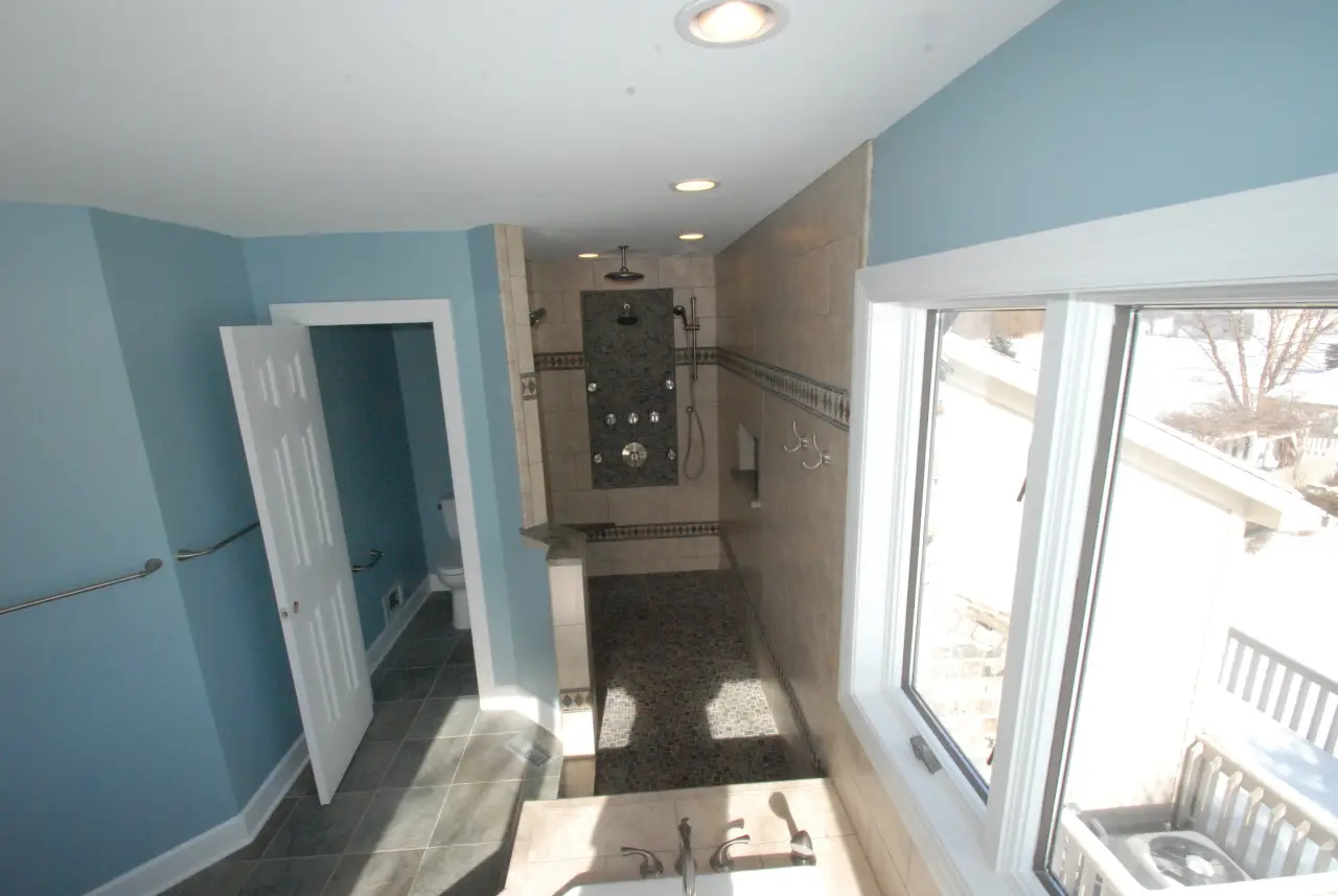 A spacious bathroom featuring blue walls, a large shower area with multiple fixtures, and natural light from a window.