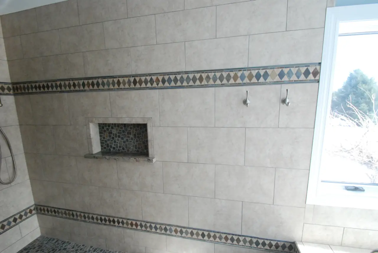 A tiled bathroom wall featuring a decorative border, a recessed shelf, and two hooks, with a window providing natural light.