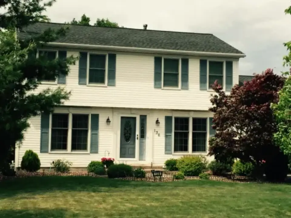 Two-story white house with blue shutters, front door, and a green lawn with bushes and trees.