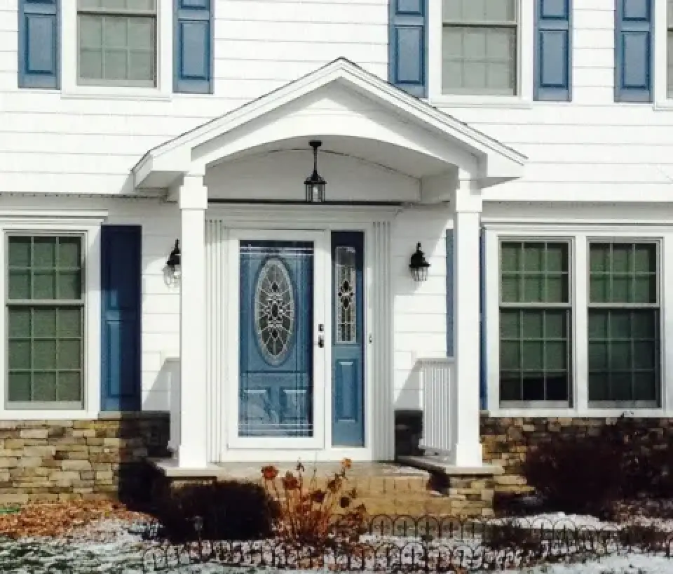 Front entrance of a white house with blue door, sidelights, columns, and some winter landscaping.