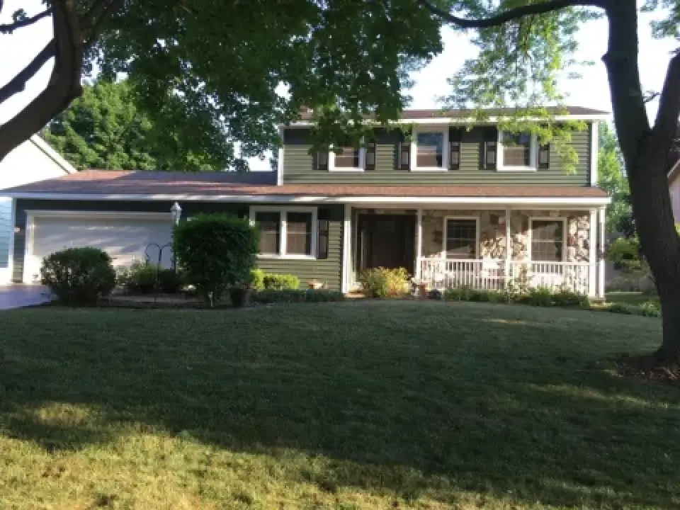 Green two-story house with white trim, front porch, and a well-kept lawn shaded by trees.