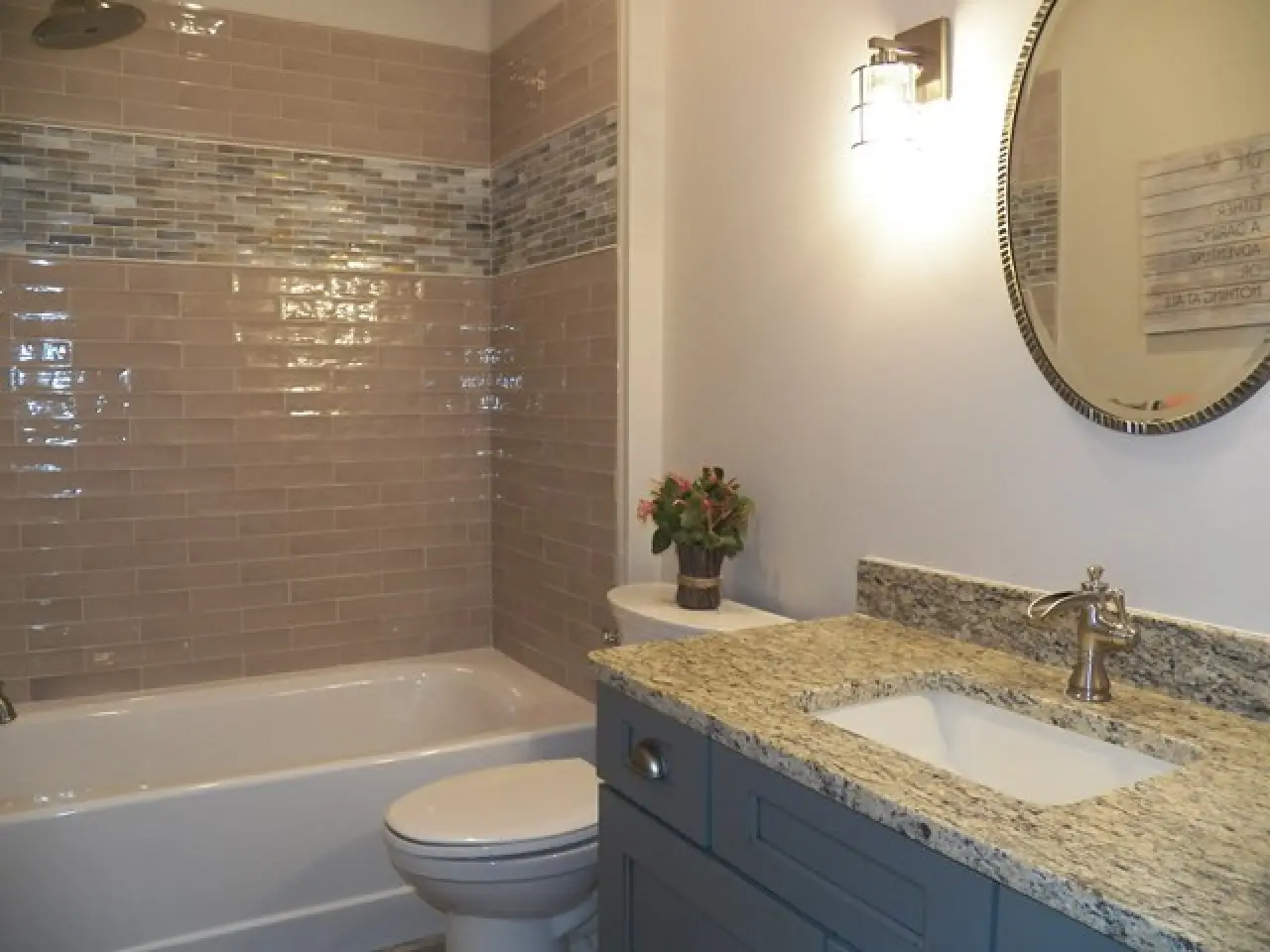 A modern bathroom featuring a bathtub, beige tiled walls, a granite countertop, and a round mirror with a light fixture.