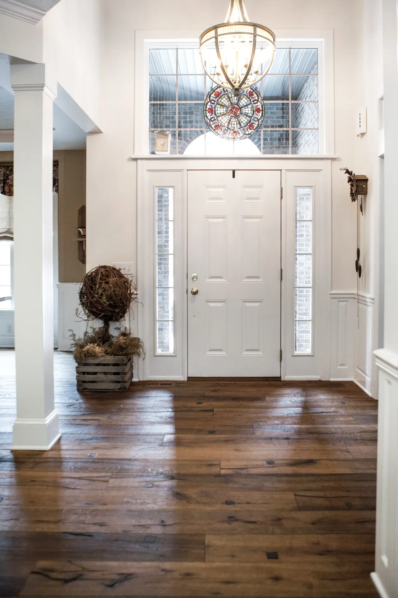 A bright entryway featuring a white door, large windows, a chandelier, and wooden flooring, with decorative elements like a plant in a crate.