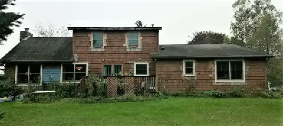 A weathered two-story house with brown siding, large windows, and a grassy yard in front.
