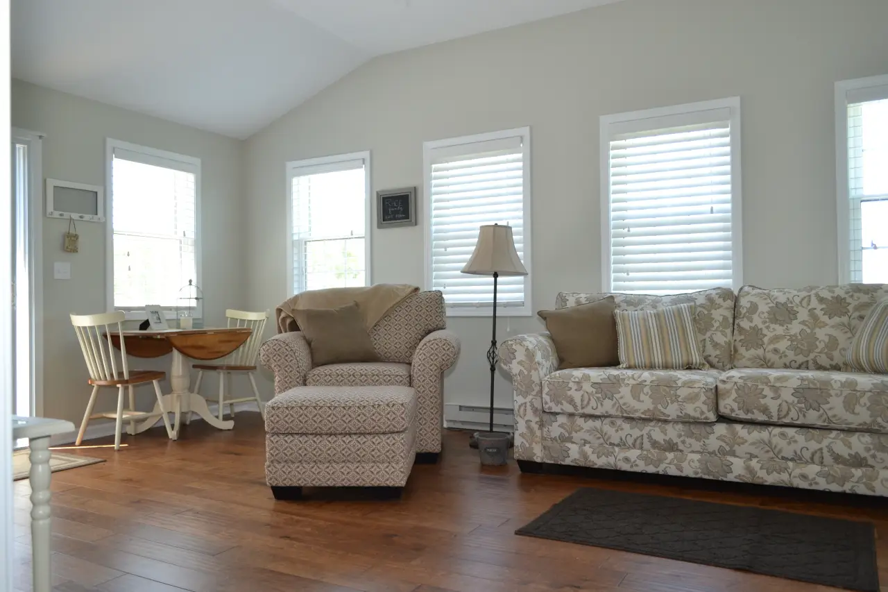 A cozy living room featuring a patterned sofa, a comfortable armchair, a small dining table, and bright windows with blinds.