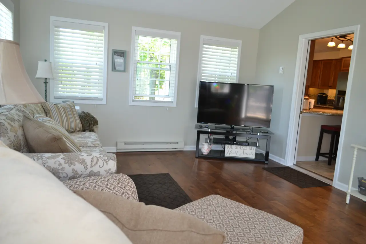 A cozy living room featuring a sofa, a TV on a stand, and large windows with blinds, leading to a kitchen area.