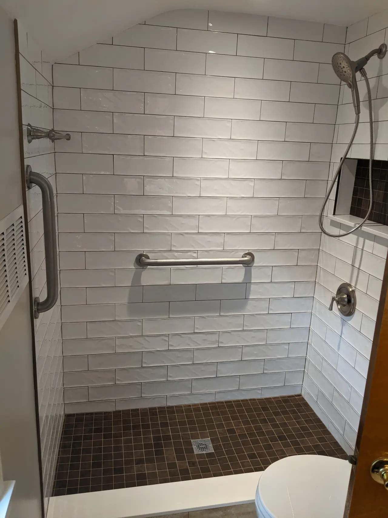 A modern shower with white tiled walls, a brown tiled floor, grab bars, and a handheld showerhead.