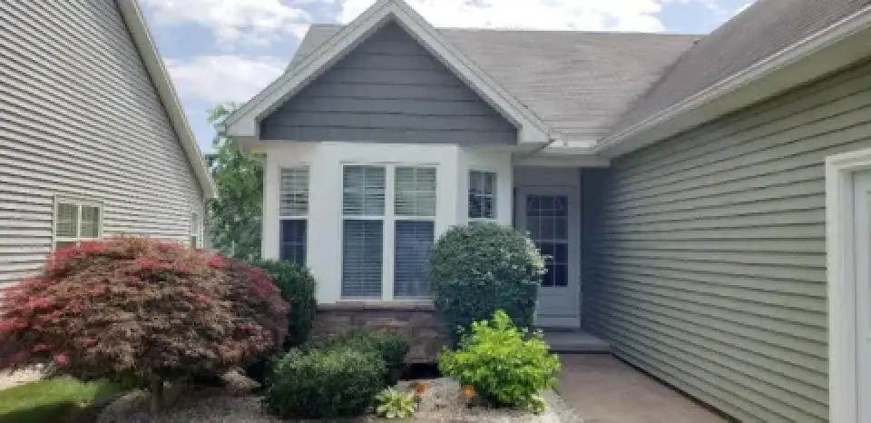 Single-story house exterior with green siding, front bushes, and a white-trimmed window.