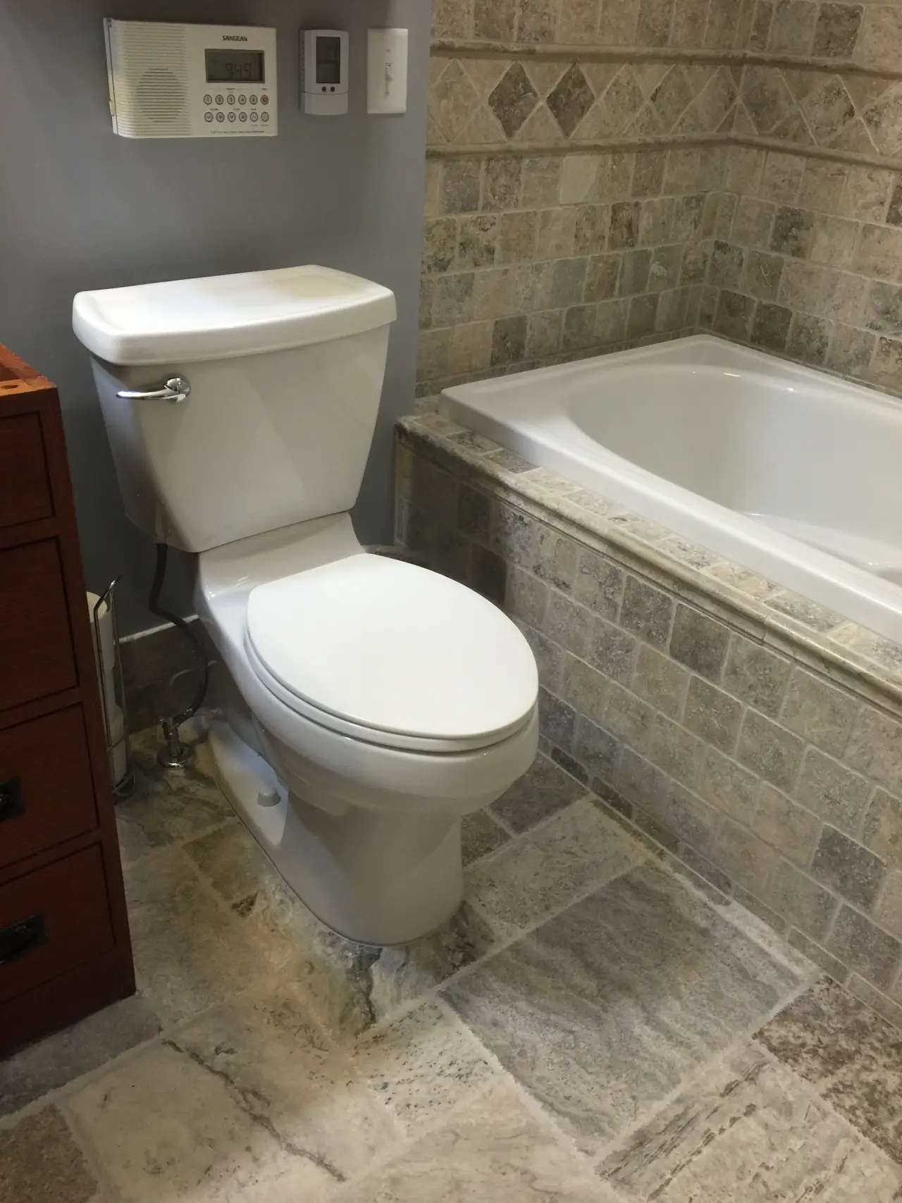 A bathroom featuring a white toilet, a bathtub, and stone tile flooring. There are also wall-mounted controls and a wooden cabinet.