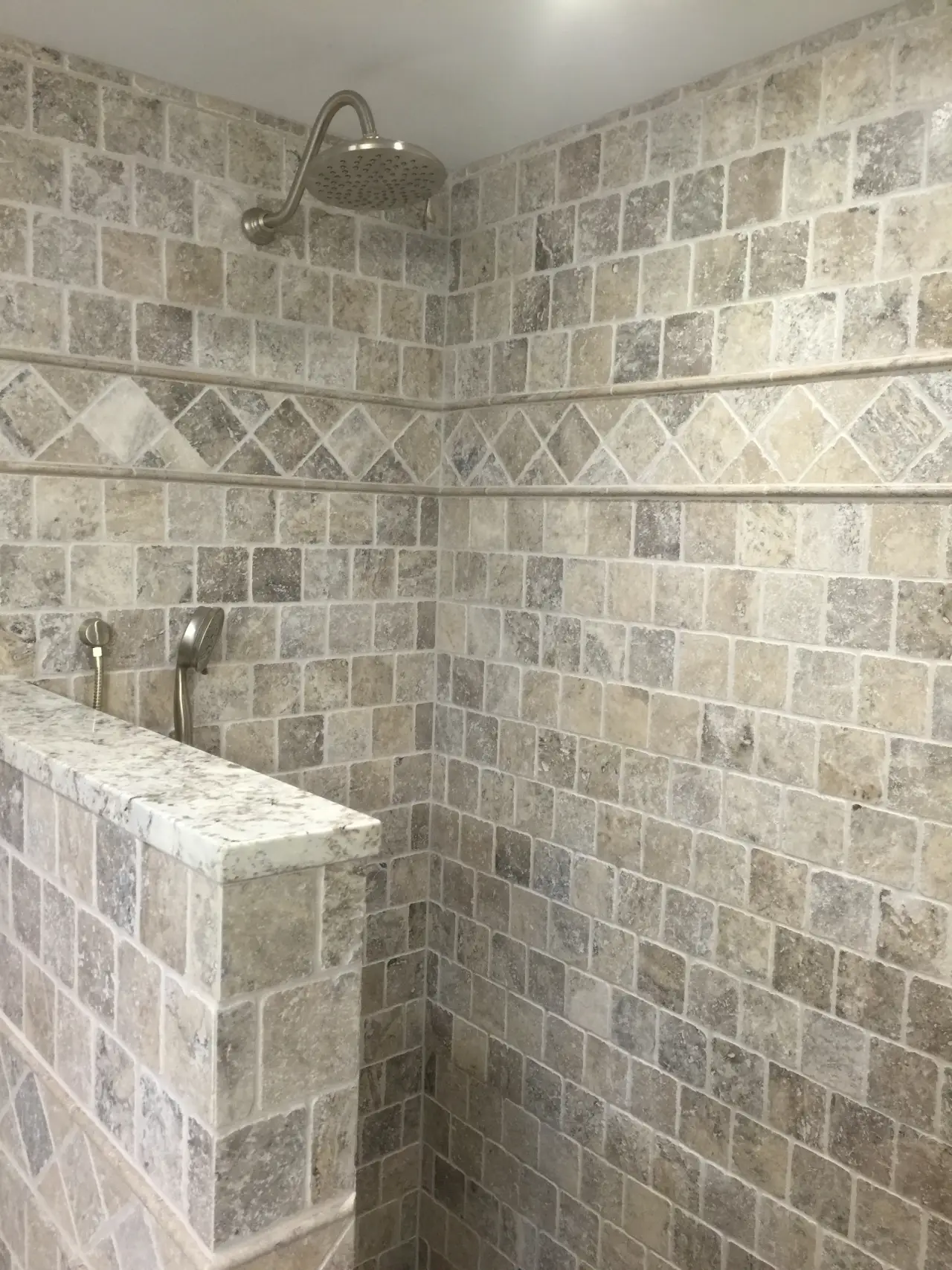A modern shower with textured stone tiles in neutral tones, featuring a wall-mounted showerhead and two faucet controls.