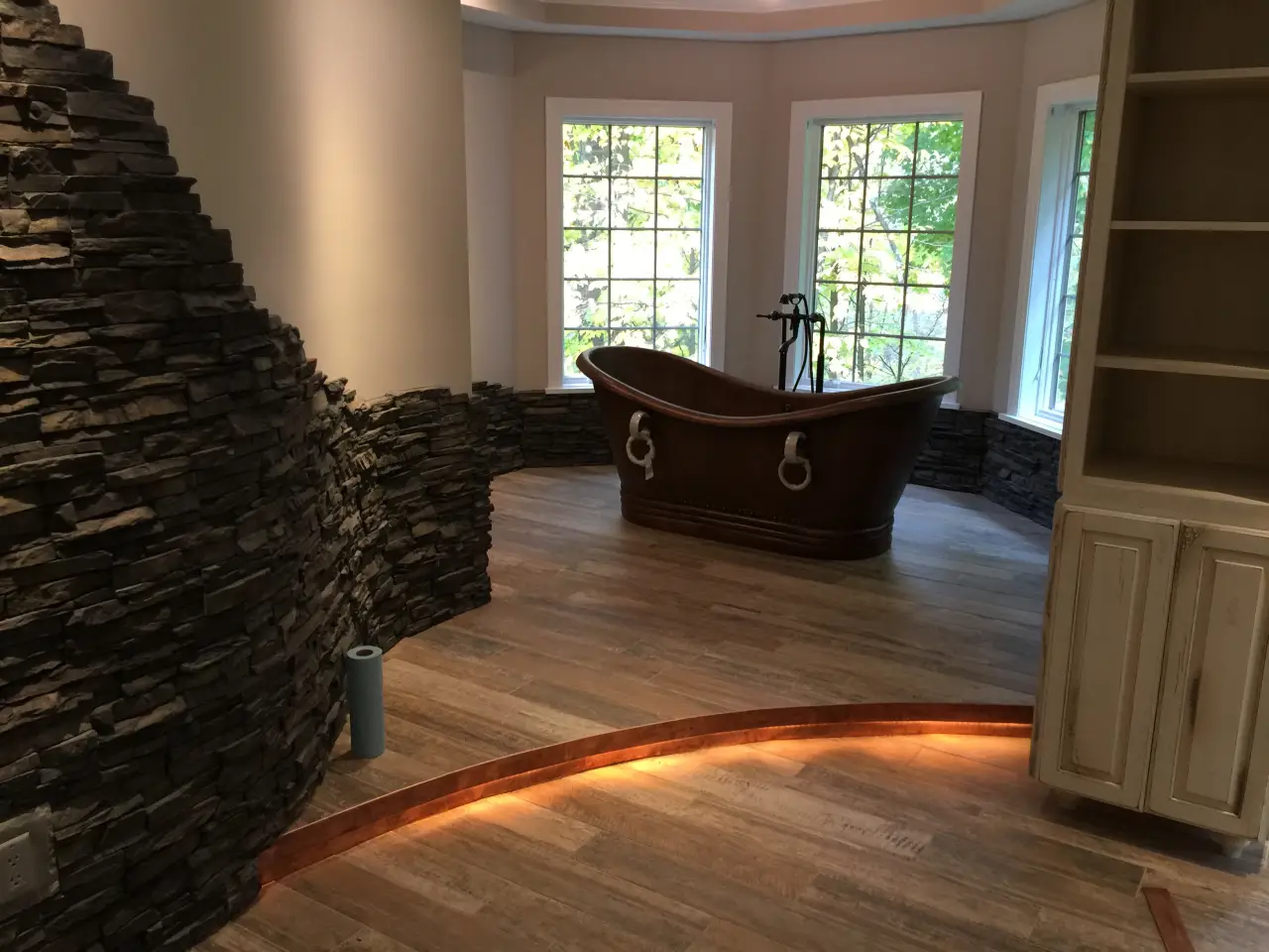 A stylish bathroom featuring a wooden bathtub, stone wall accents, large windows, and warm lighting on the floor.
