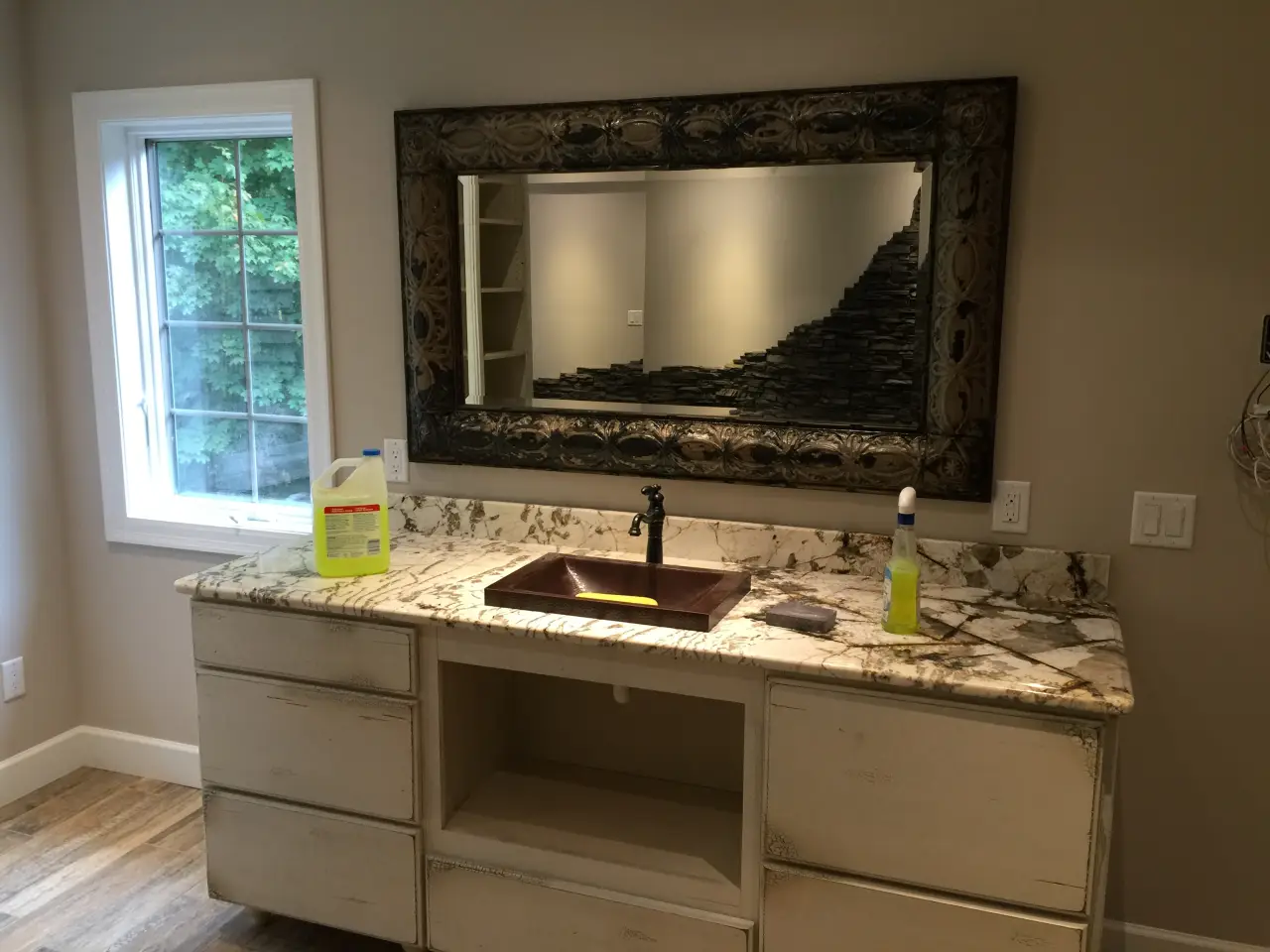 A bathroom vanity with a marble countertop, a brown sink, and a large decorative mirror. Cleaning supplies are on the counter.
