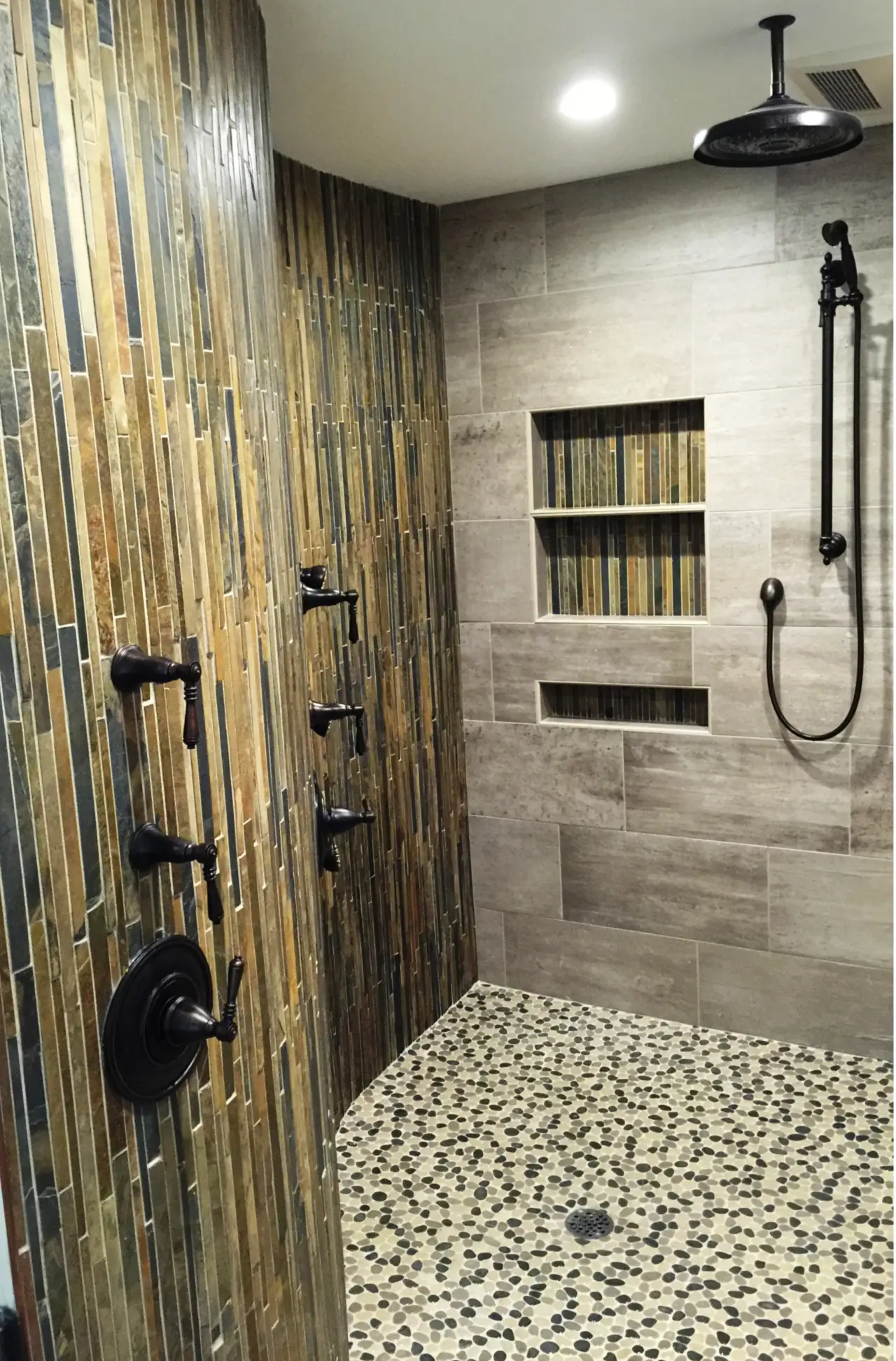 A modern shower with textured stone walls, a pebble floor, and black fixtures. Features a built-in shelf for toiletries.