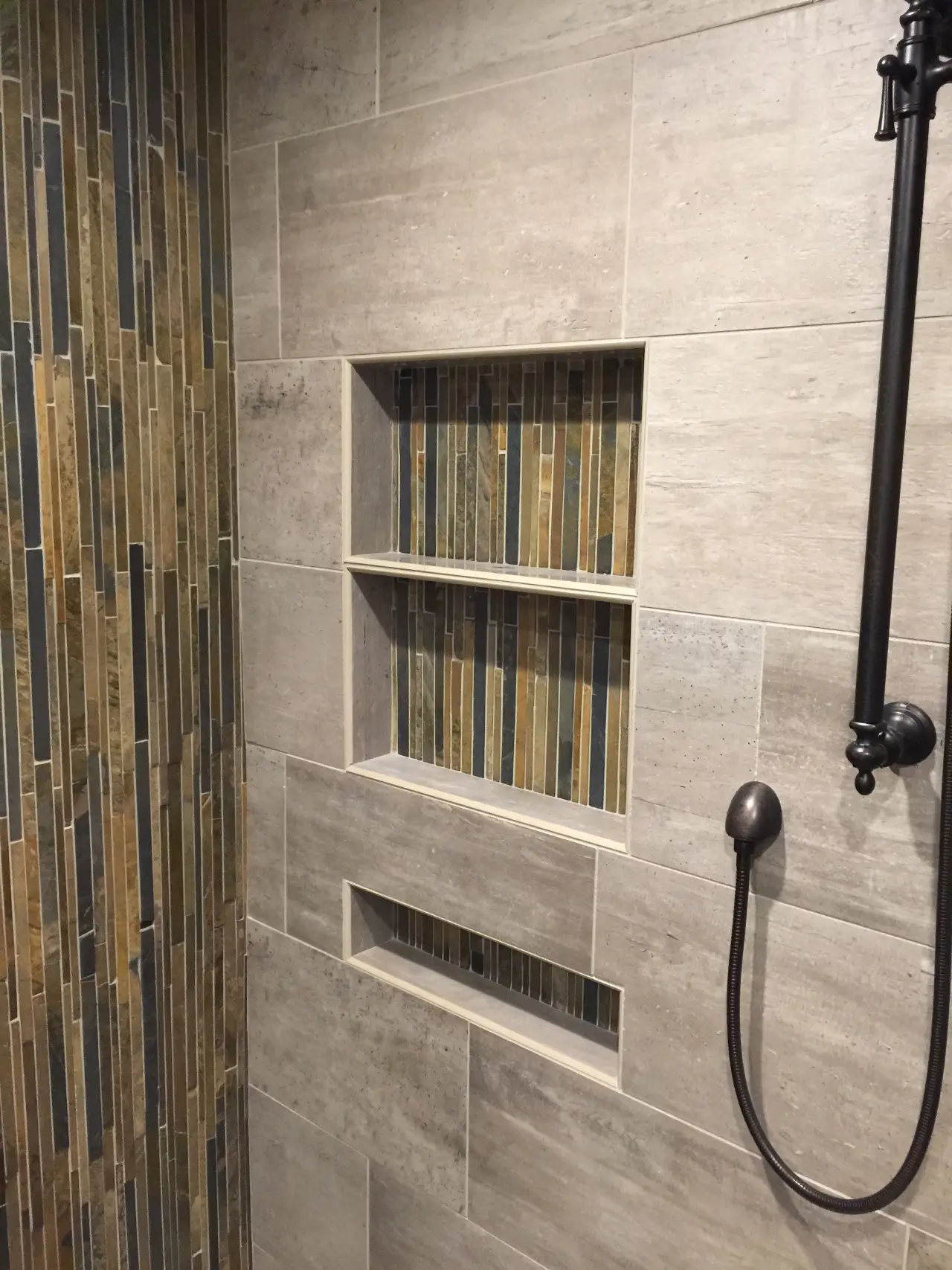 A modern shower wall featuring textured tiles in gray and earthy tones, with built-in shelves for storage.