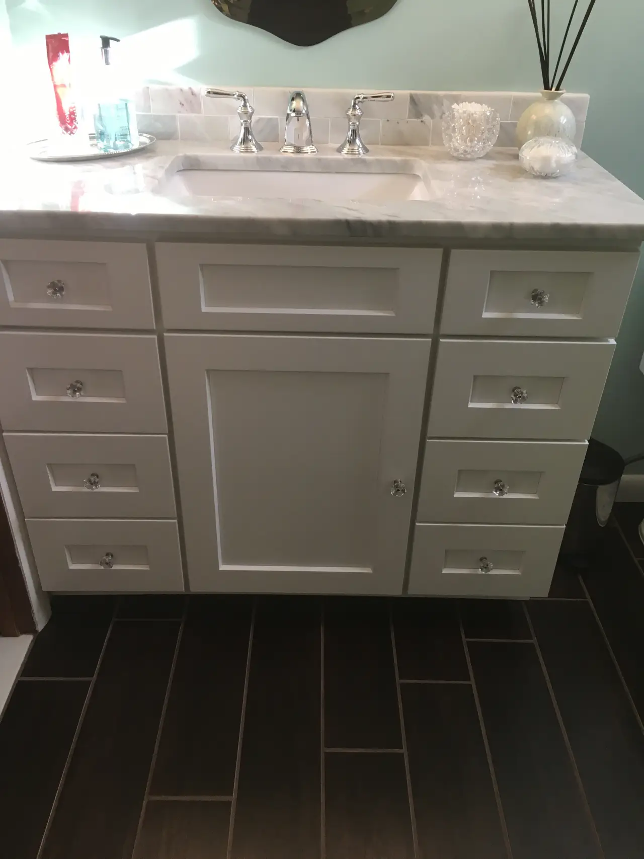 A modern bathroom vanity with a marble countertop, silver faucets, and multiple white drawers with crystal knobs. Dark tiled floor.