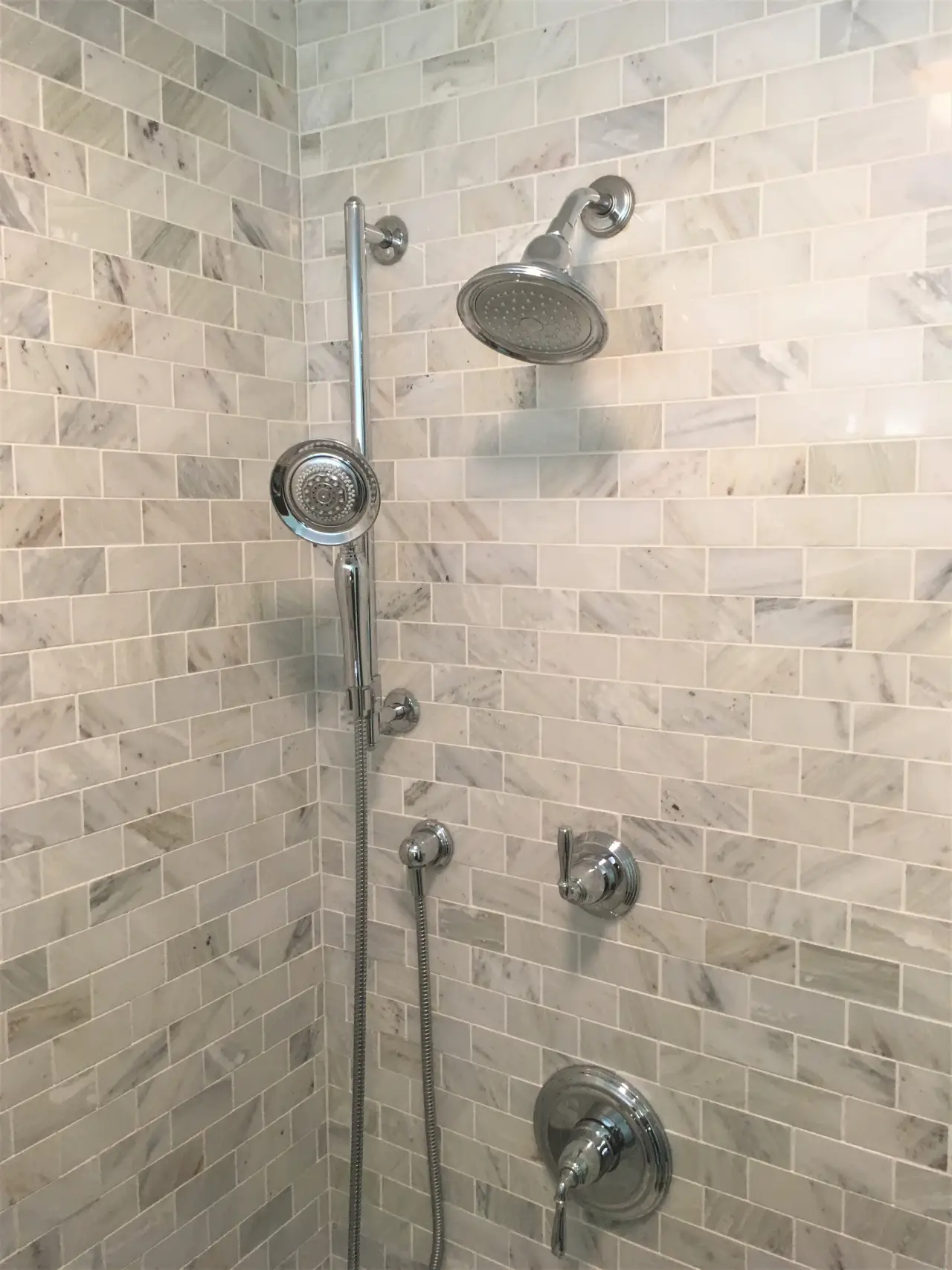 A modern shower with two chrome showerheads and a handheld sprayer, set against a light marble tile wall.