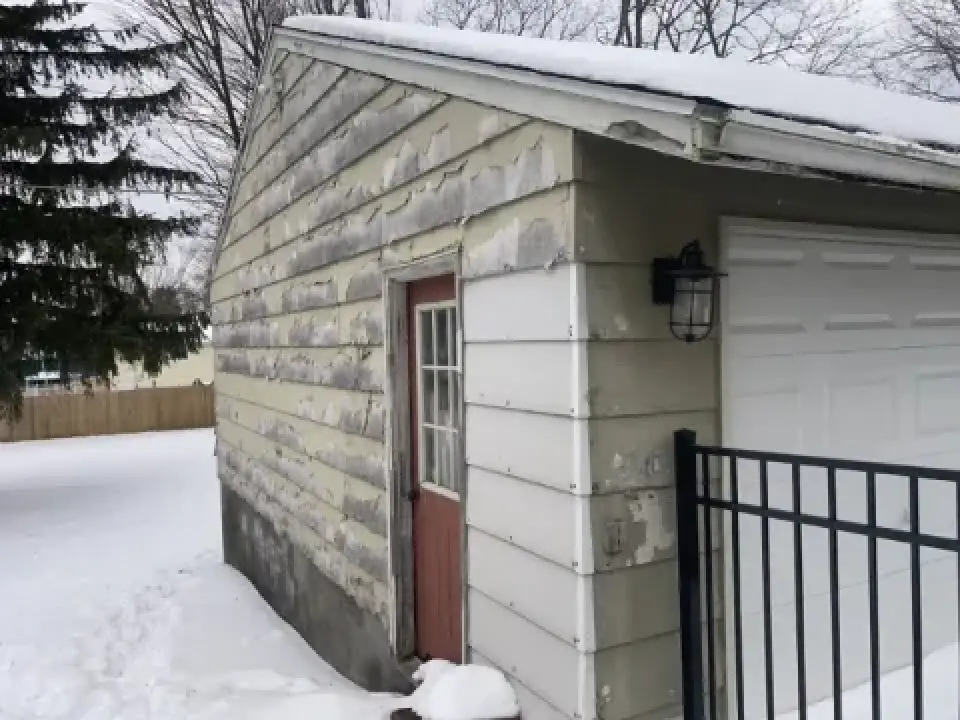 A weathered garage with peeling paint and snow on the ground and roof.
