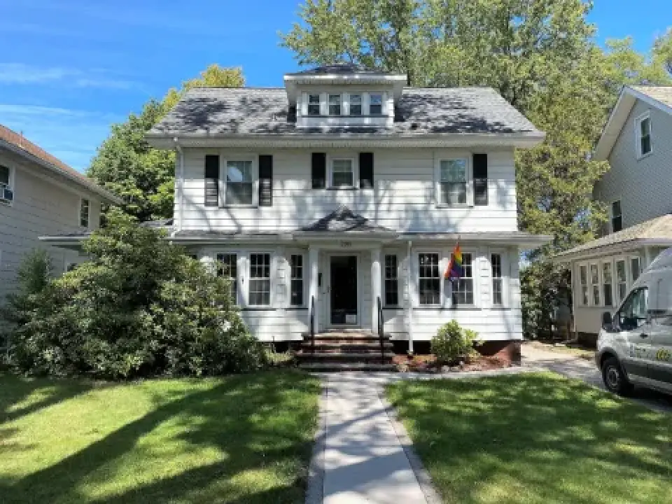 Two-story white house with black shutters, rainbow flag, and a white van parked on the right.